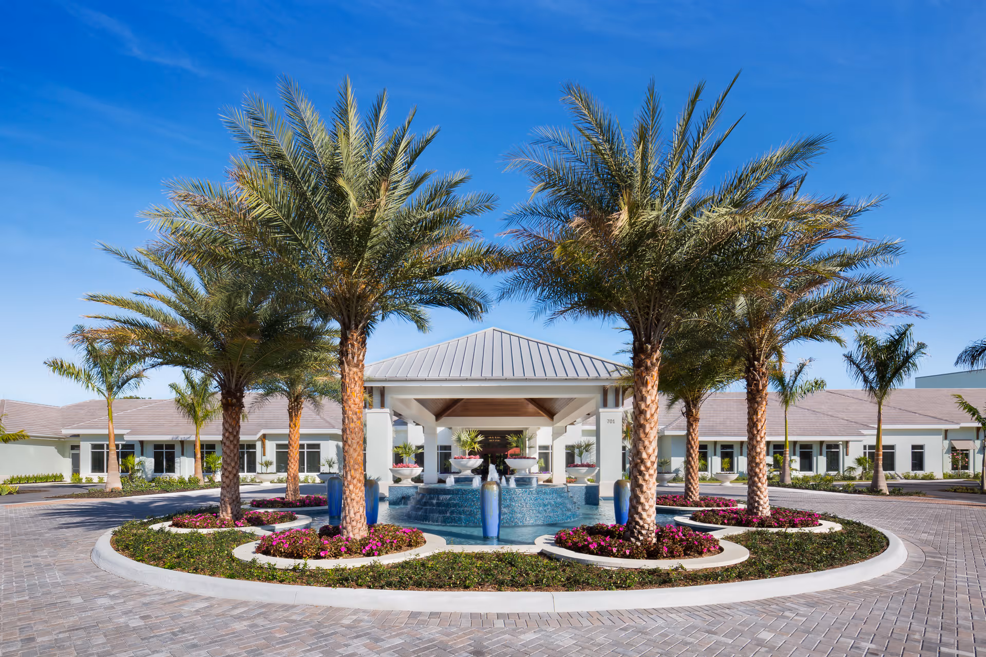 Front exterior view of Vi at Bentley Village featuring a circular driveway with palm trees and flower beds surrounding a central water fountain under a covered entrance with a clear blue sky background.