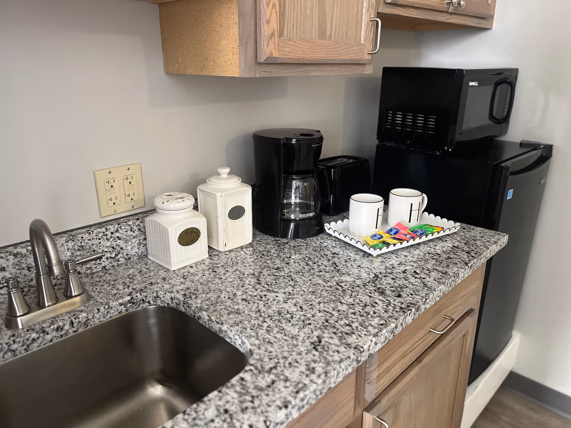 A small kitchen countertop with a stainless steel sink, a coffee maker, a toaster, two white ceramic containers labeled Tea and Coffee, and a tray holding two white mugs and several packets of sweetener. Above the countertop are wooden cabinets, and to the right is a black mini refrigerator with a microwave on top.