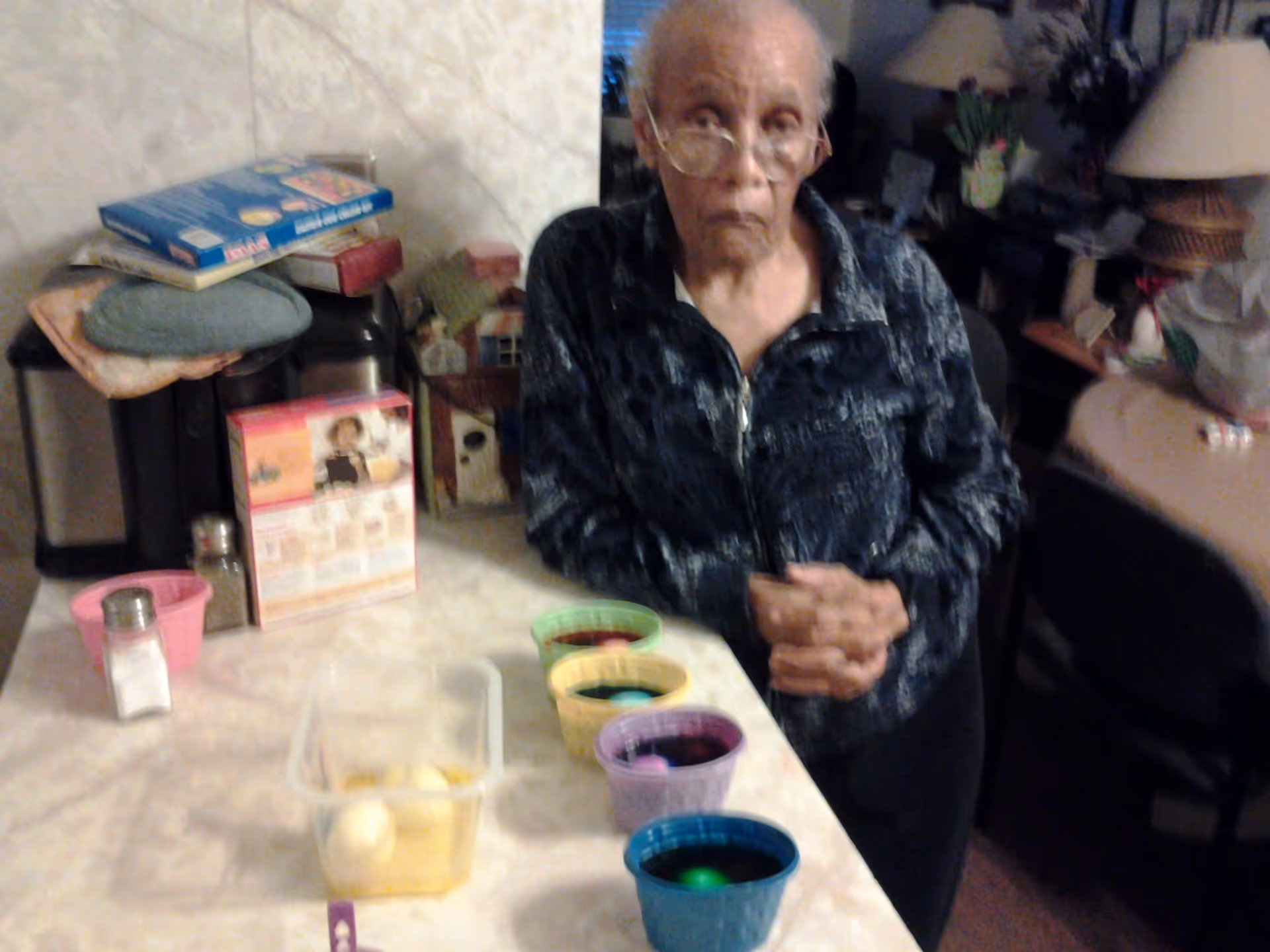 An elderly person wearing glasses and a dark patterned jacket stands at a kitchen counter with several colorful cups filled with dyed water and eggs, presumably for egg dyeing. The kitchen counter also has a container with white eggs, a salt shaker, and some boxes and kitchen items in the background.