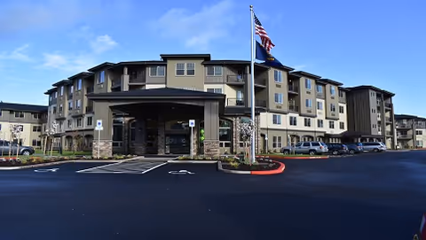Exterior view of a multi-story senior living facility building with a covered entrance, several parked cars, and two flagpoles displaying the American flag and another flag. The sky is clear with some clouds.