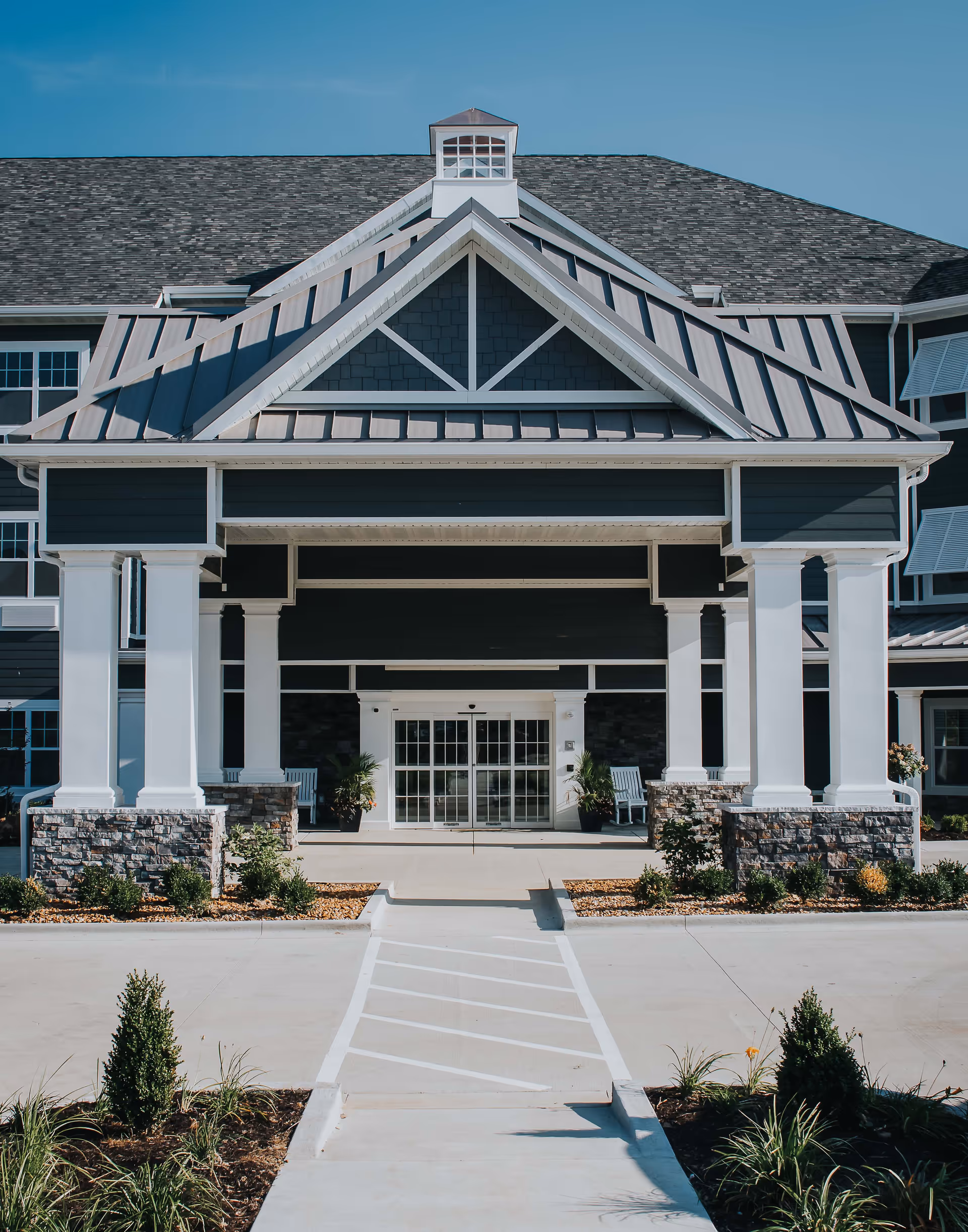Front entrance of a senior living facility with a covered driveway supported by white columns and stone bases, glass double doors, and landscaped plants along the walkway under a clear blue sky.