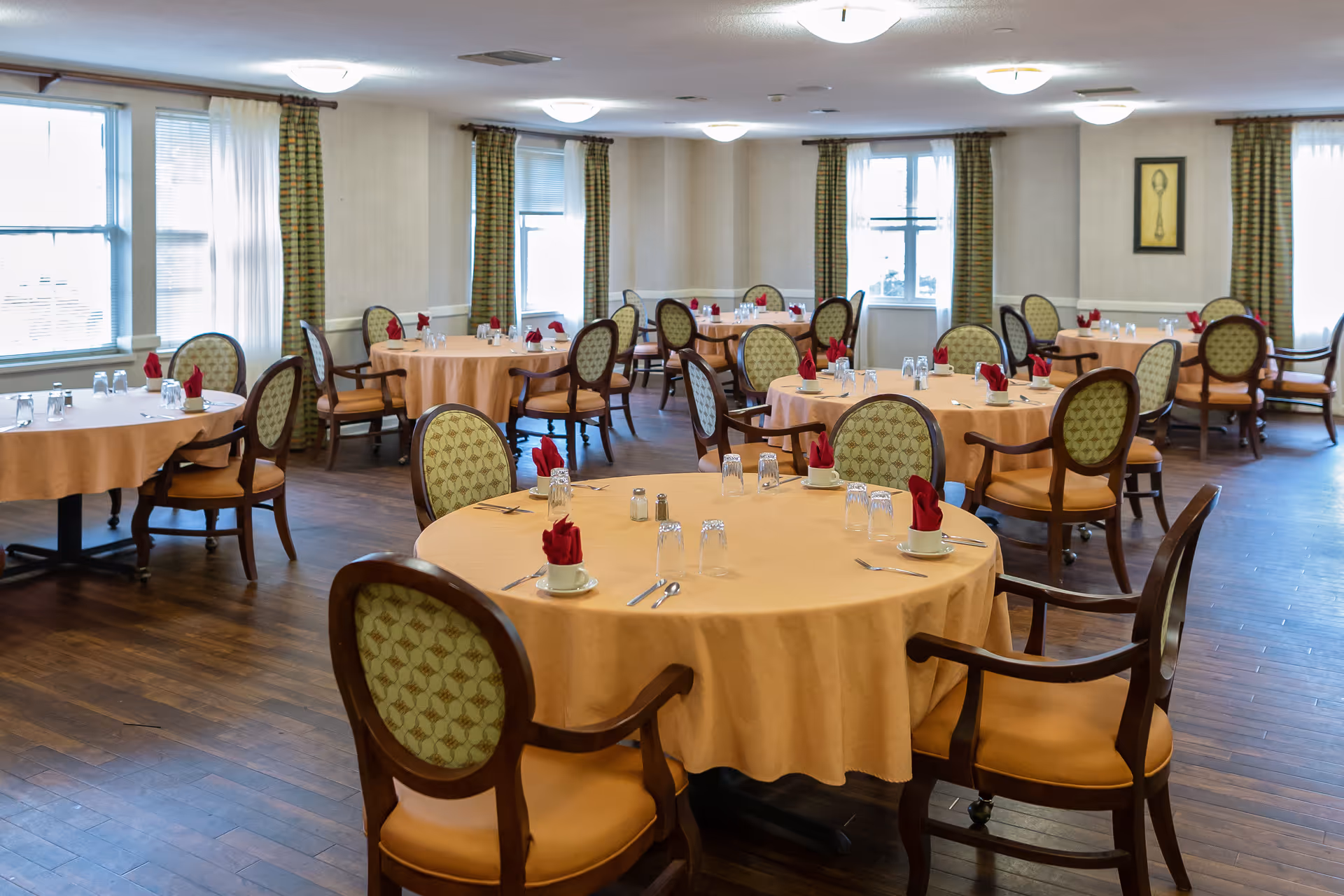 A dining room with multiple round tables covered with beige tablecloths, each set with cups, glasses, silverware, and red folded napkins. The room has wooden floors, patterned chairs with armrests, large windows with curtains, and soft ceiling lights.