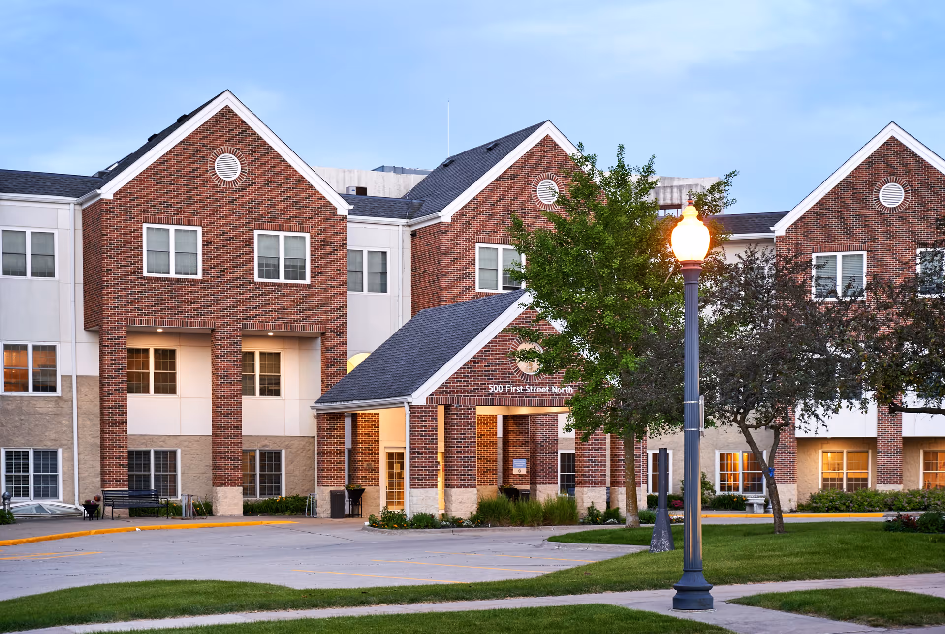 Brick-faced senior living building with a covered entrance, illuminated streetlamp, trees, and a manicured lawn.