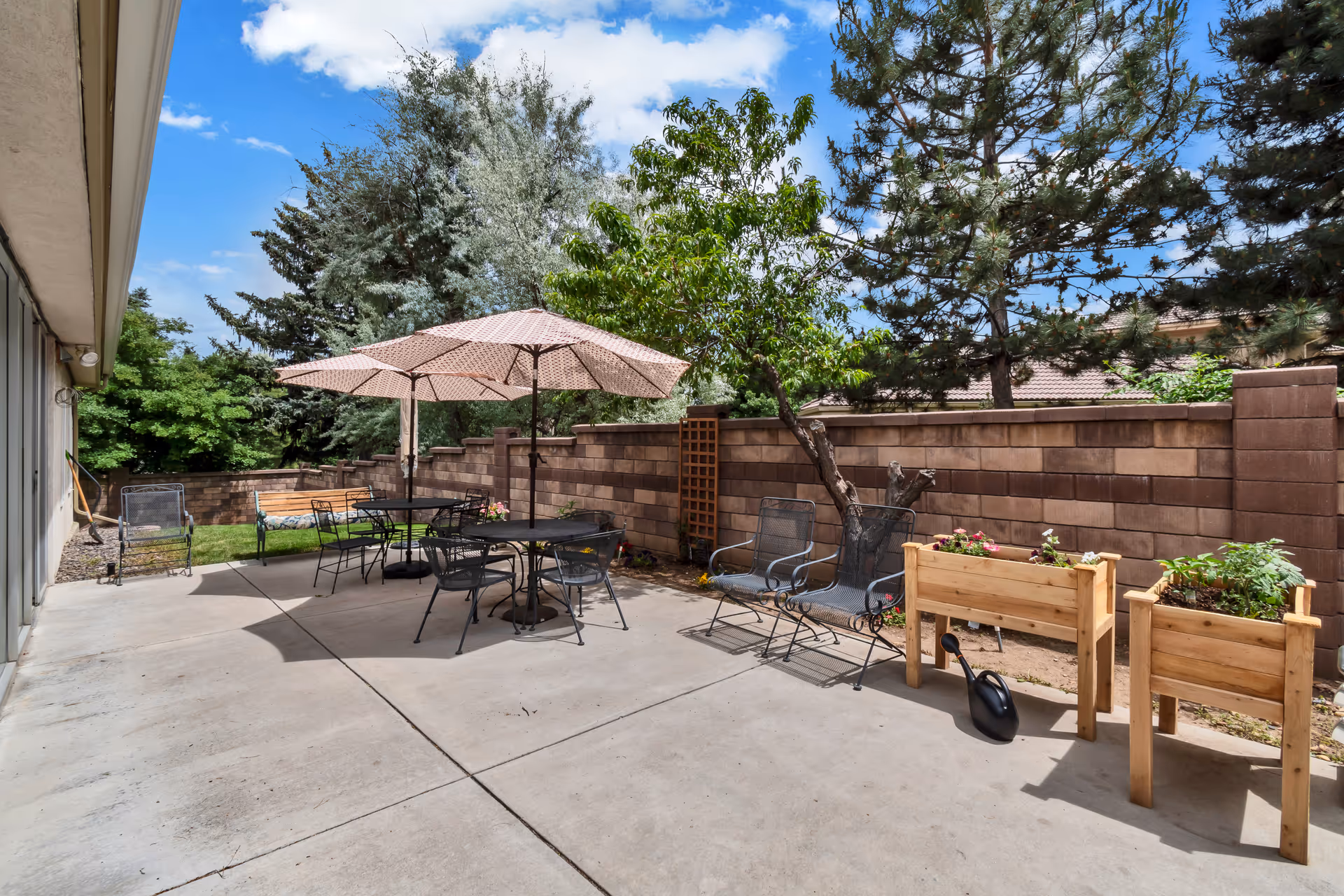 Outdoor patio area with concrete flooring, featuring two round tables with umbrellas and metal chairs. There are additional metal chairs and wooden planters with plants along a brick wall. Trees and greenery are visible beyond the wall under a partly cloudy blue sky.
