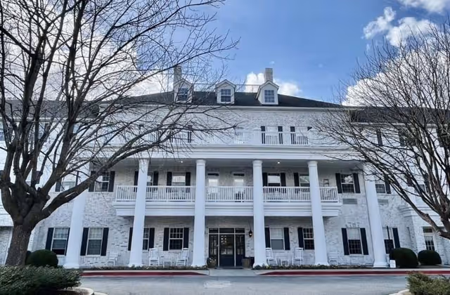 Front exterior view of a large white brick building with black shutters and multiple windows. The building has a covered porch supported by white columns, with rocking chairs arranged on the porch. Two leafless trees frame the building on either side under a partly cloudy blue sky.