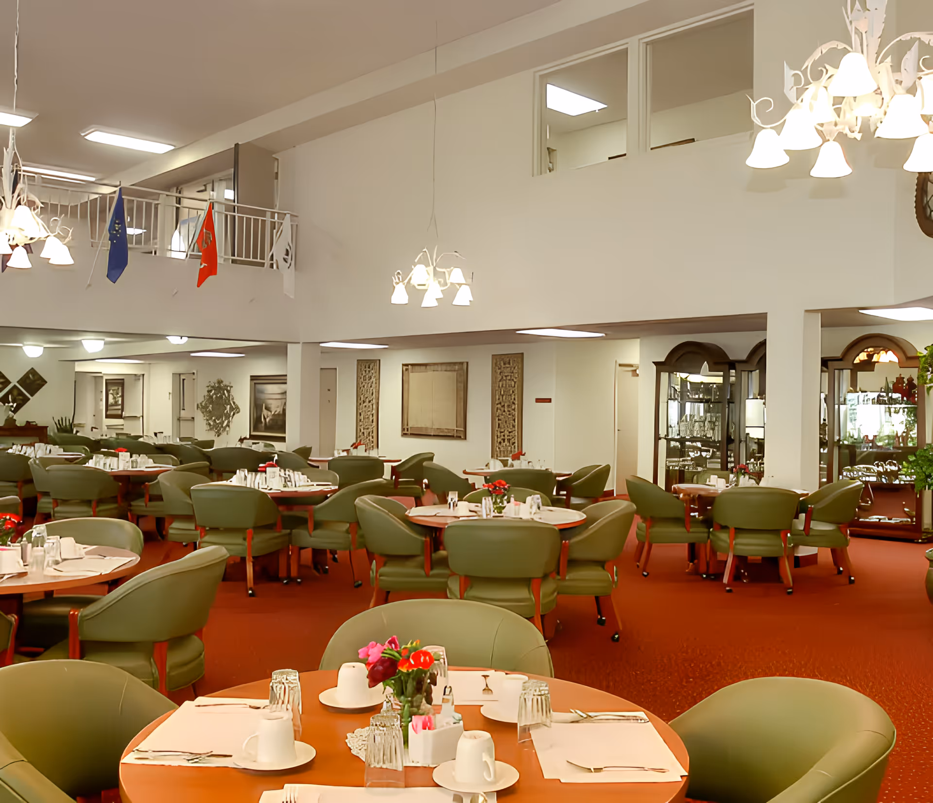 A spacious dining room with multiple round tables set with placemats, cups, glasses, and silverware. The room features green cushioned chairs, red carpet flooring, white walls, and decorative elements including flags hanging from a balcony railing and framed artwork on the walls. There are chandeliers hanging from the ceiling providing warm lighting.