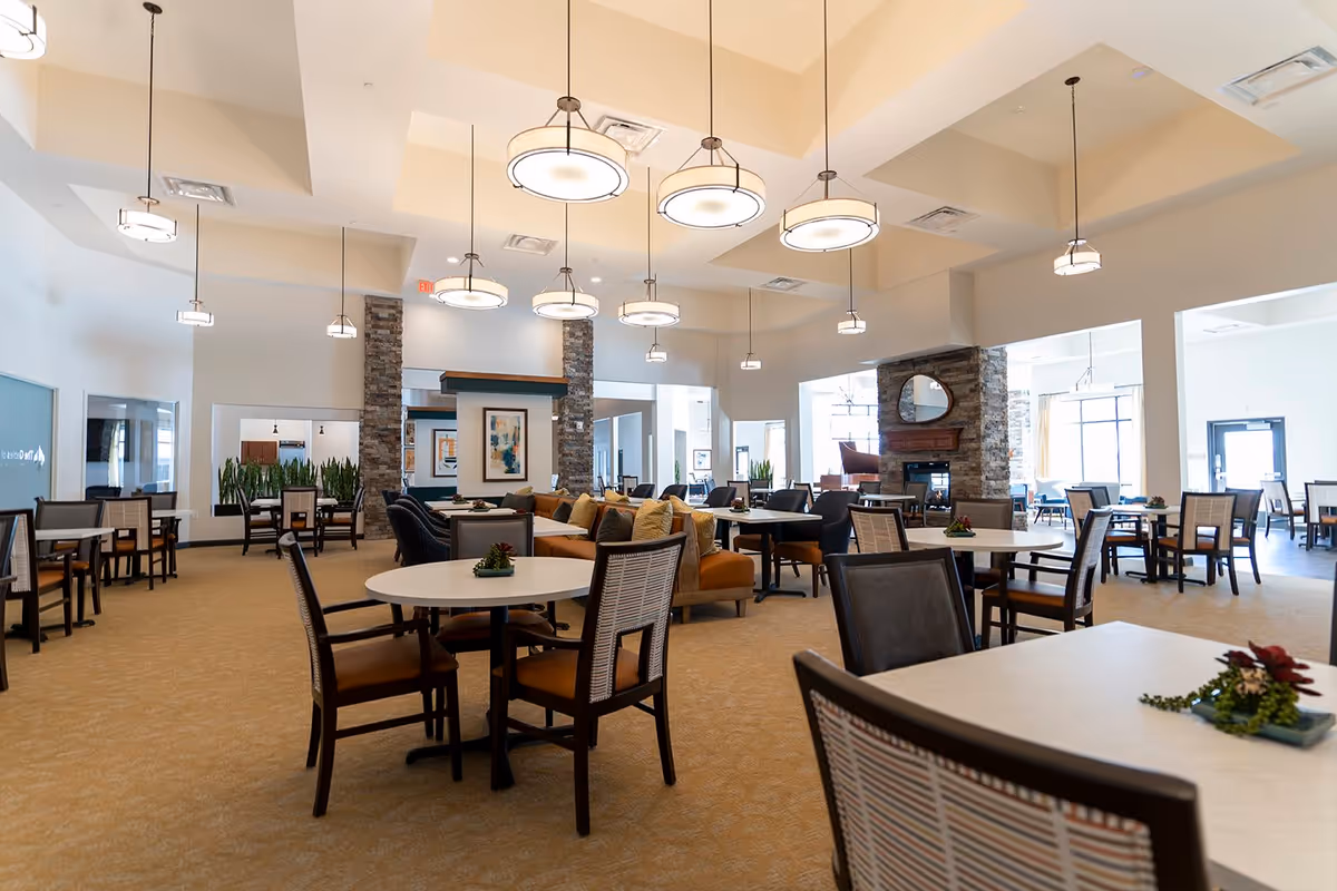 A spacious and well-lit dining area in a senior living facility with multiple round and rectangular tables surrounded by chairs. The room features high ceilings with modern hanging light fixtures, stone pillars, and a cozy fireplace with a mirror above it. There are decorative plants on the tables and large windows allowing natural light to fill the space.
