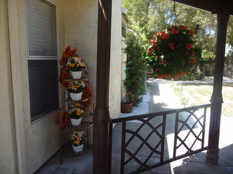 View from a covered porch showing a decorative metal plant stand with four white pots of colorful flowers next to a window. A hanging basket with red flowers is suspended from the porch ceiling. Beyond the porch is a pathway leading to a garden area with trees and greenery.