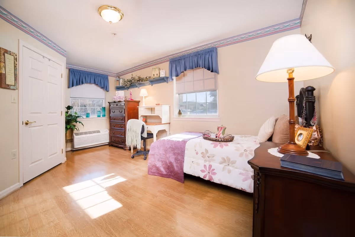 Sunny furnished bedroom with a single bed, nightstand and lamp, desk and dresser, and windows with blue valances.