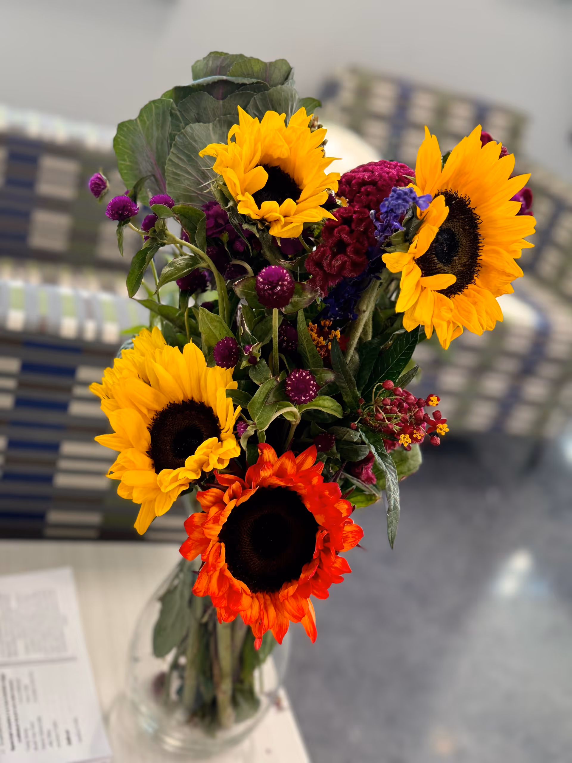 A vibrant bouquet of flowers in a clear glass vase, featuring bright yellow sunflowers, a red sunflower, and various purple and green foliage, placed on a white table with blurred patterned chairs in the background.
