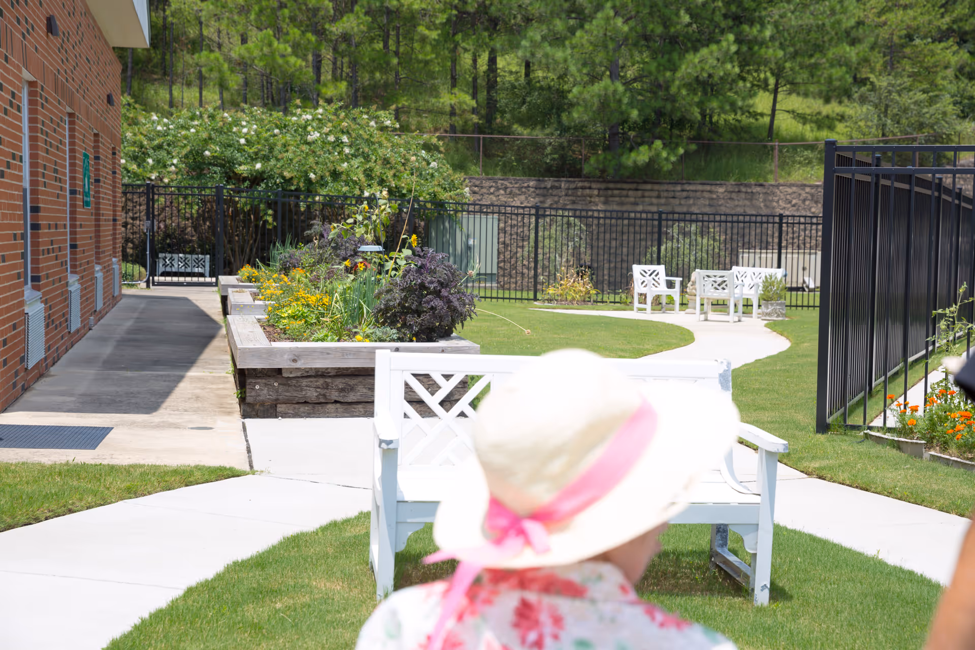 Outdoor garden area at a senior living facility with white benches, a paved walkway, raised flower beds with various plants and flowers, a brick building on the left, and a person wearing a white hat with a pink ribbon in the foreground.