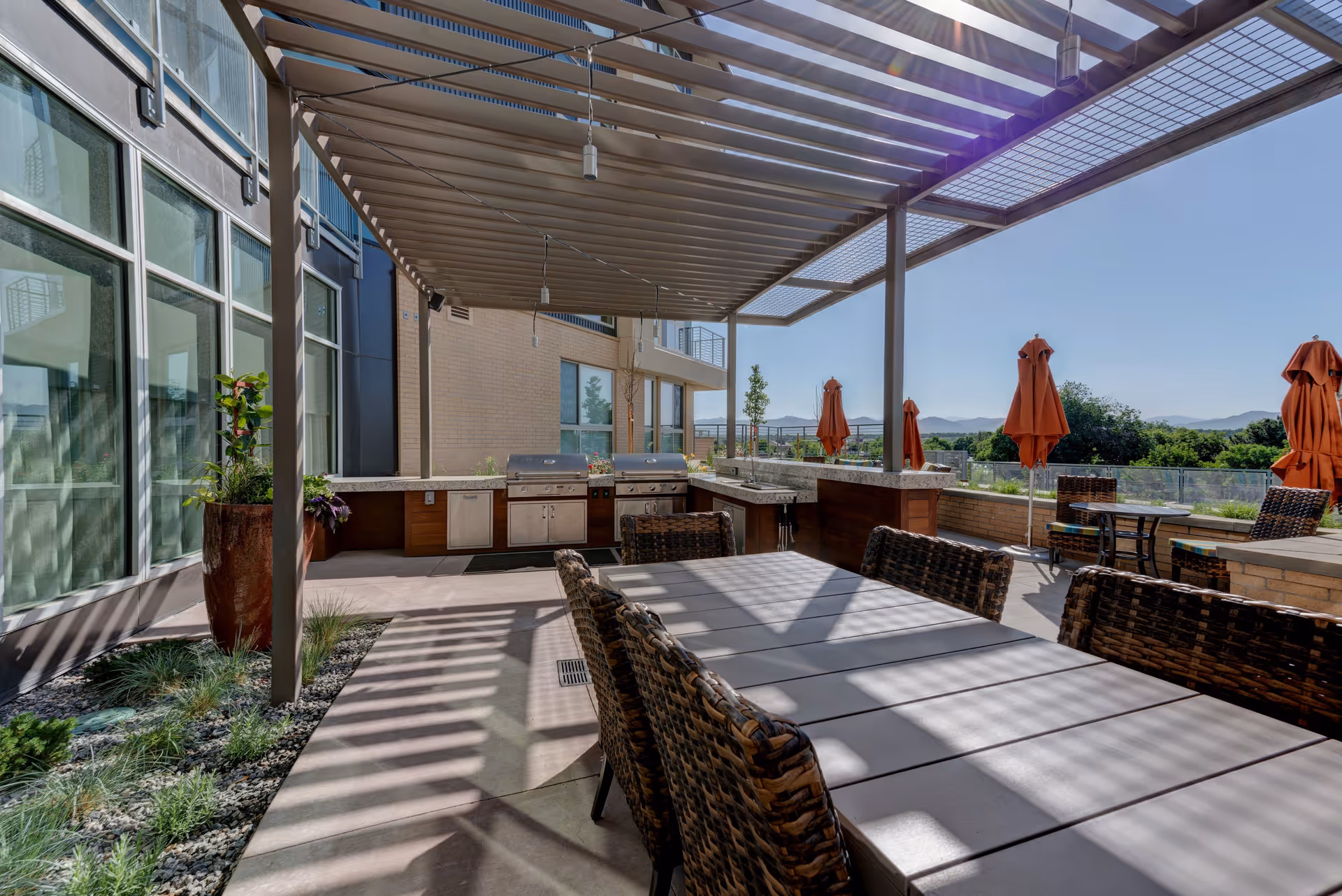 Outdoor patio area with a large dining table surrounded by wicker chairs under a pergola. There are two stainless steel grills and a countertop with a sink in the background. Several orange umbrellas are closed and positioned around smaller tables. The area overlooks greenery and distant mountains under a clear blue sky.