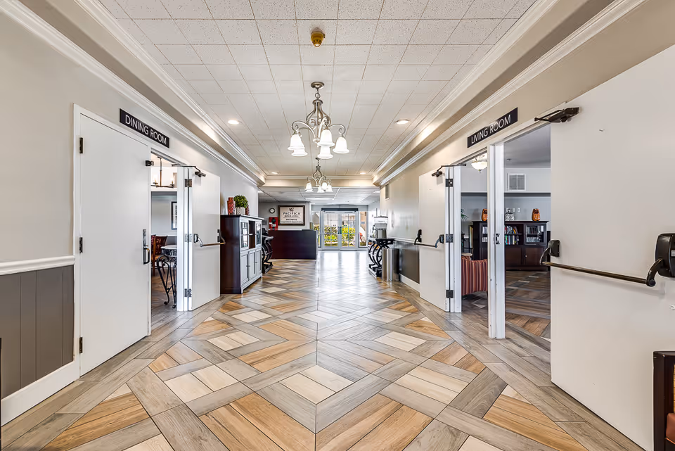 Bright interior hallway in a senior living facility with doors labeled Dining Room and Living Room and patterned wood flooring.