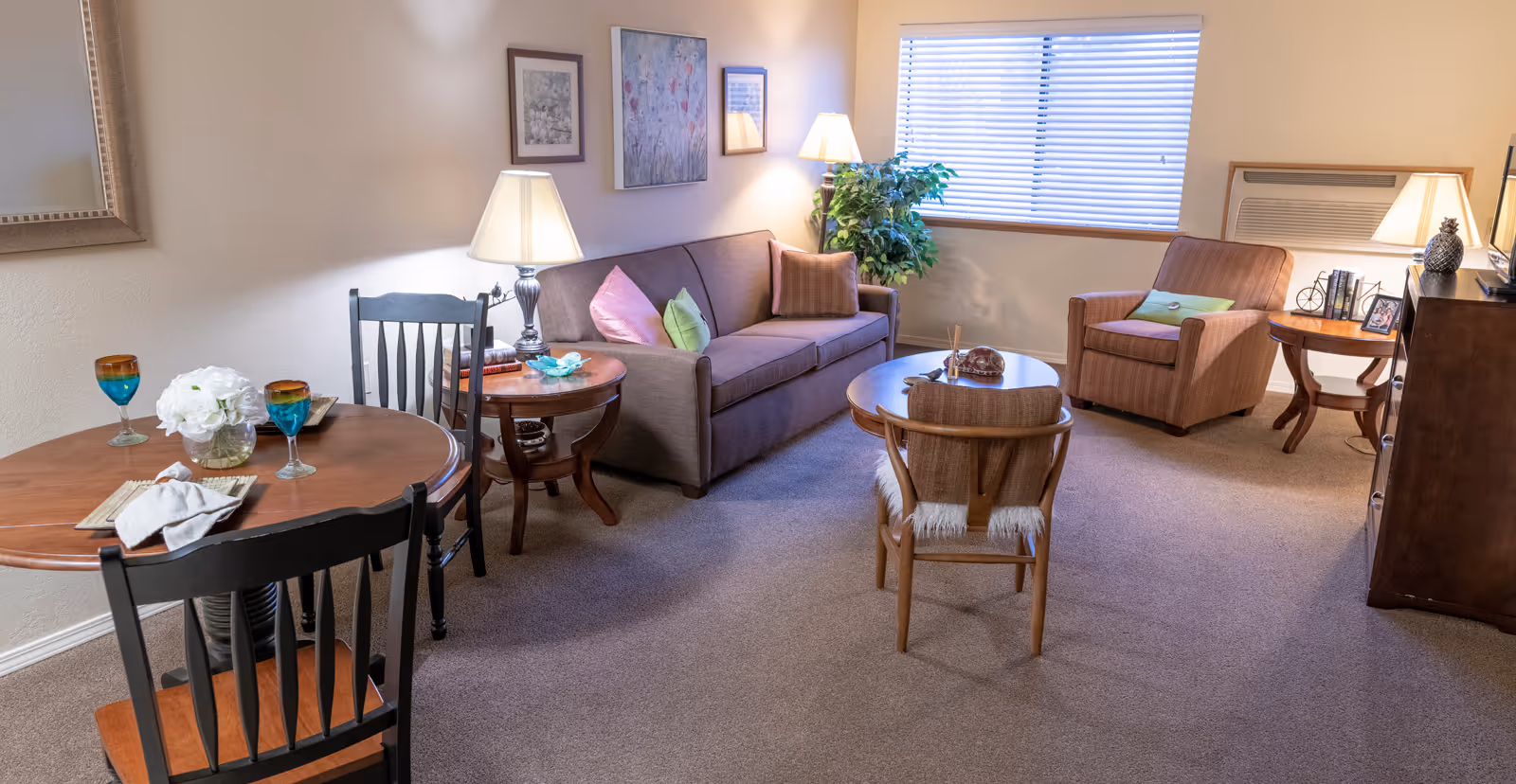 Well-lit living room with a sofa, armchairs, a small dining table, lamps, and a window with blinds.