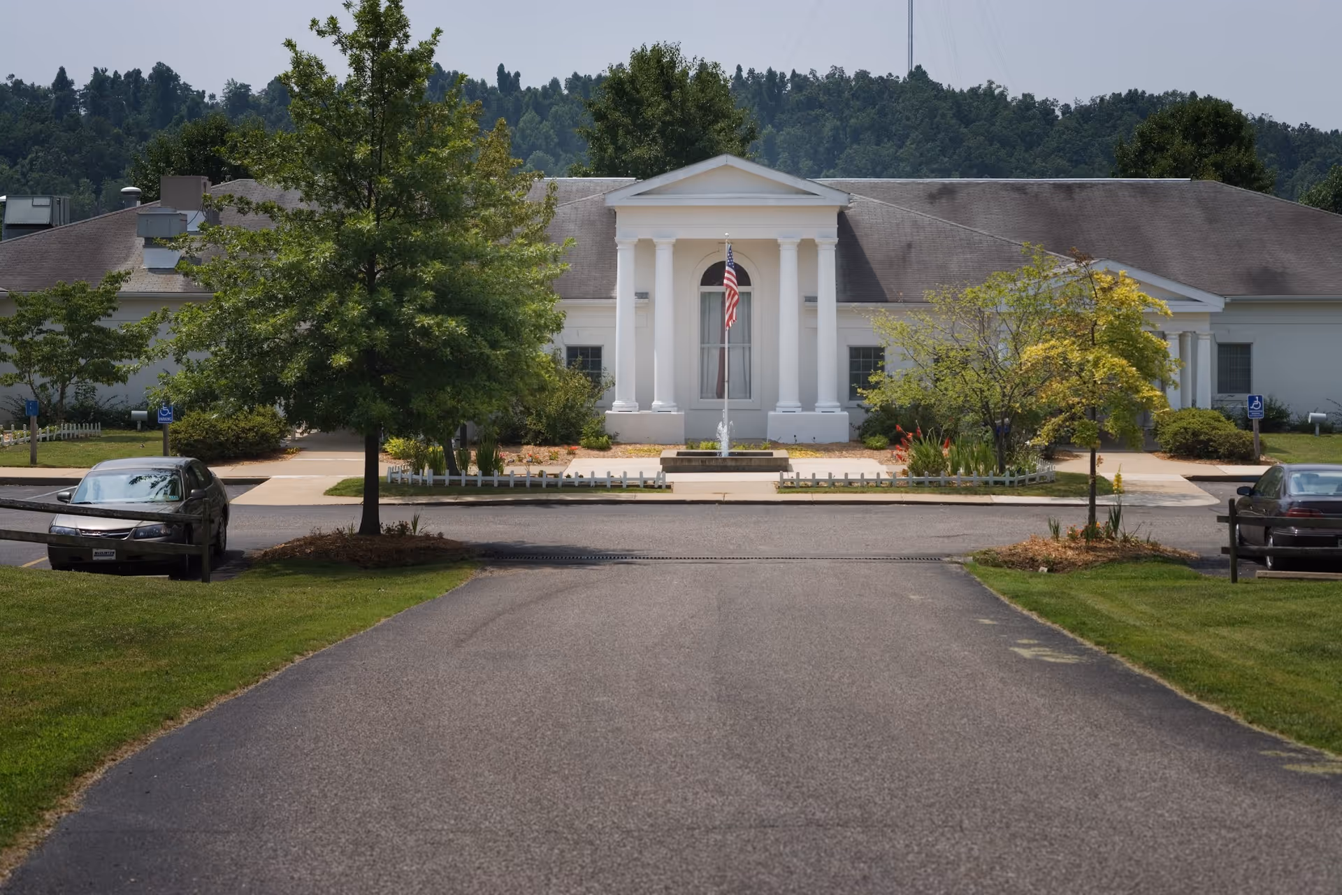 Front exterior view of a white building with classical columns and an American flag in front. The building is surrounded by trees and greenery, with a paved driveway leading up to it. Two cars are parked on either side of the driveway.