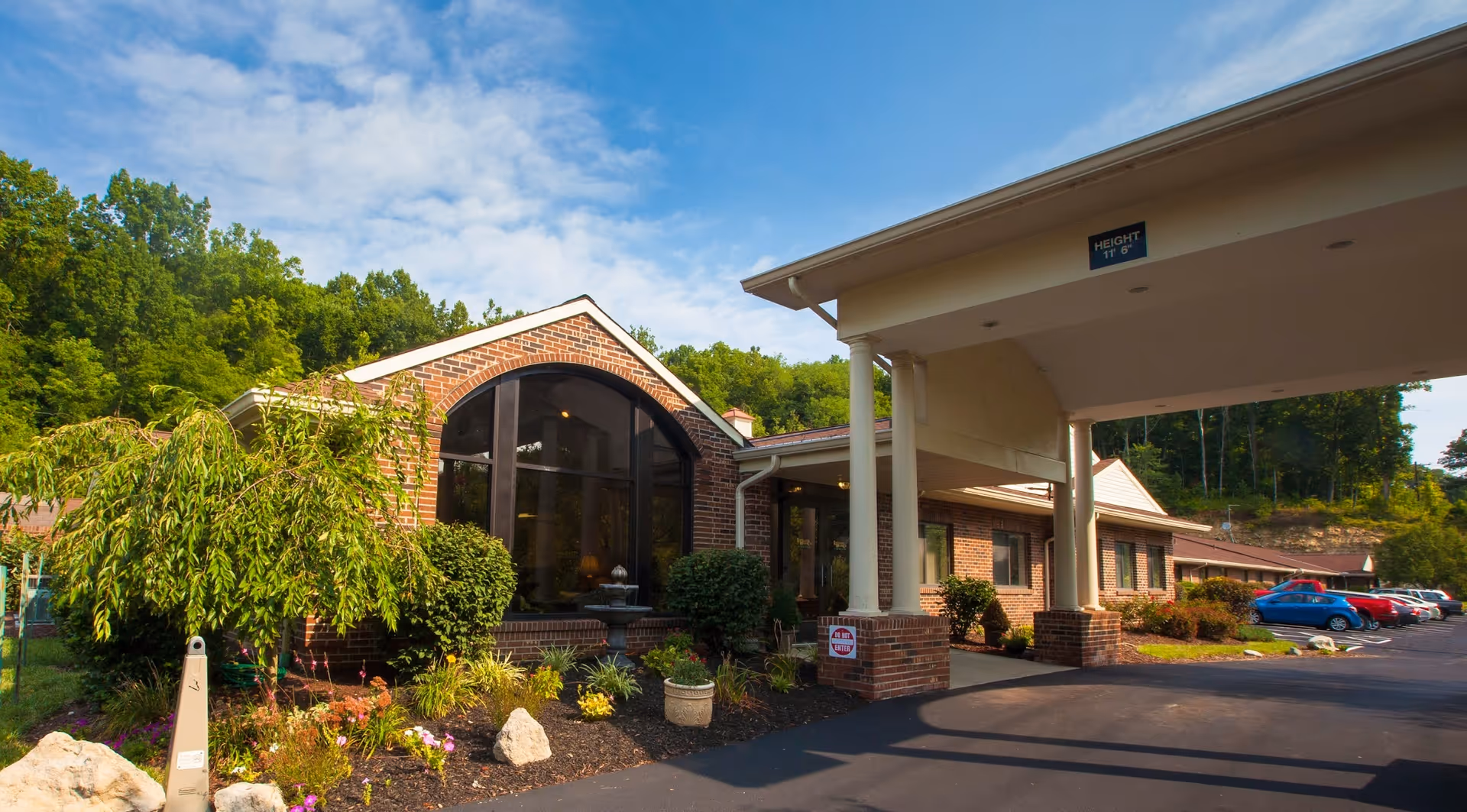 Exterior view of Loveland Health Care facility showing a brick building with large arched windows, a covered entrance supported by white columns, landscaped garden with bushes and flowers, and a parking lot with several cars under a partly cloudy sky.