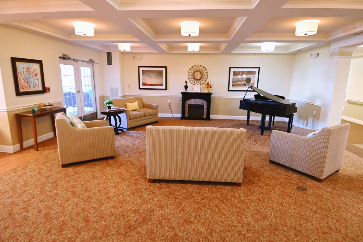 A bright communal sitting room with armchairs and sofas arranged around a decorative fireplace and a grand piano.