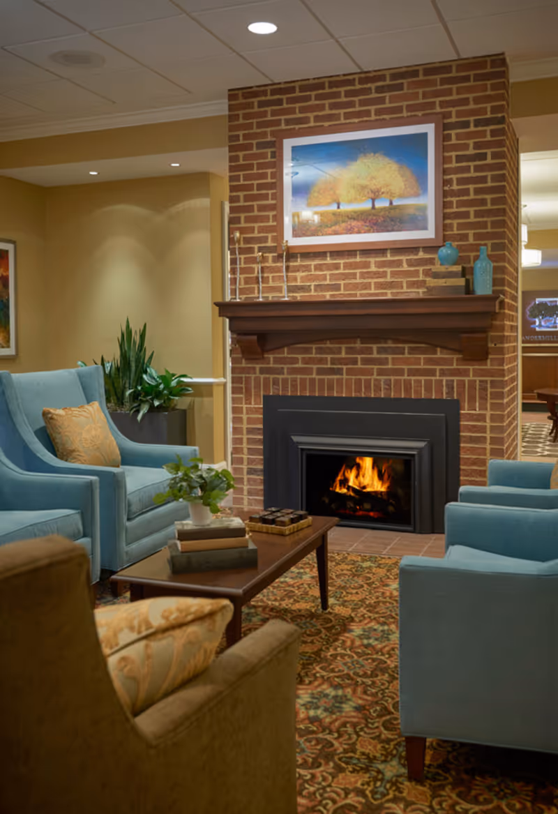 Cozy living room area with a brick fireplace featuring a burning fire. The room has comfortable blue and brown armchairs around a wooden coffee table with books and a small plant. Above the fireplace is a framed painting of trees with yellow leaves. The room is warmly lit with recessed ceiling lights and decorated with plants and vases.