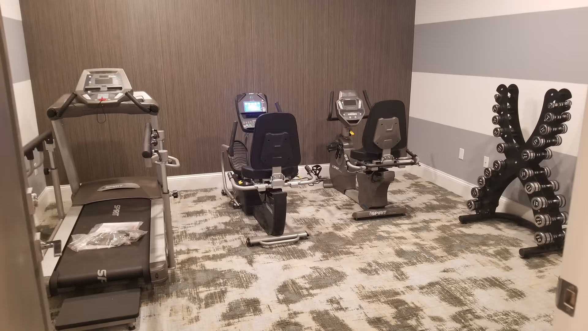 A small fitness room with a treadmill, two recumbent exercise bikes, and a rack of dumbbells against the wall. The room has patterned carpet flooring, a brown accent wall, and gray and white striped walls.