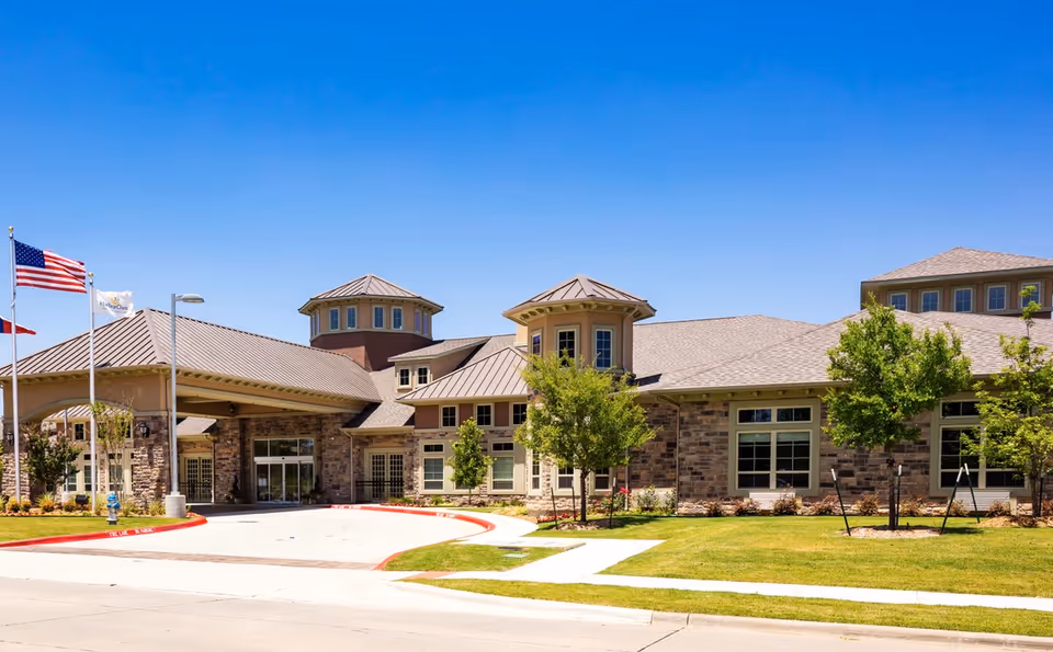 Exterior view of Village on the Park McKinney senior living facility with a clear blue sky, manicured lawn, trees, and three flagpoles displaying the American flag and other flags near the entrance.
