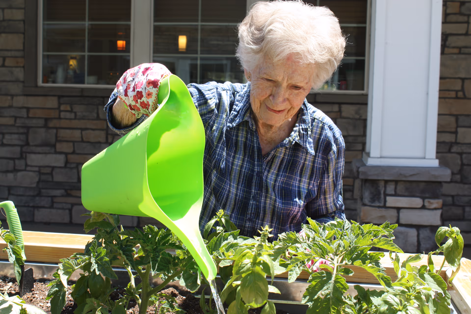 An elderly woman wearing a plaid shirt and floral gardening gloves waters plants in a raised garden bed outside a building with stone walls and large windows.