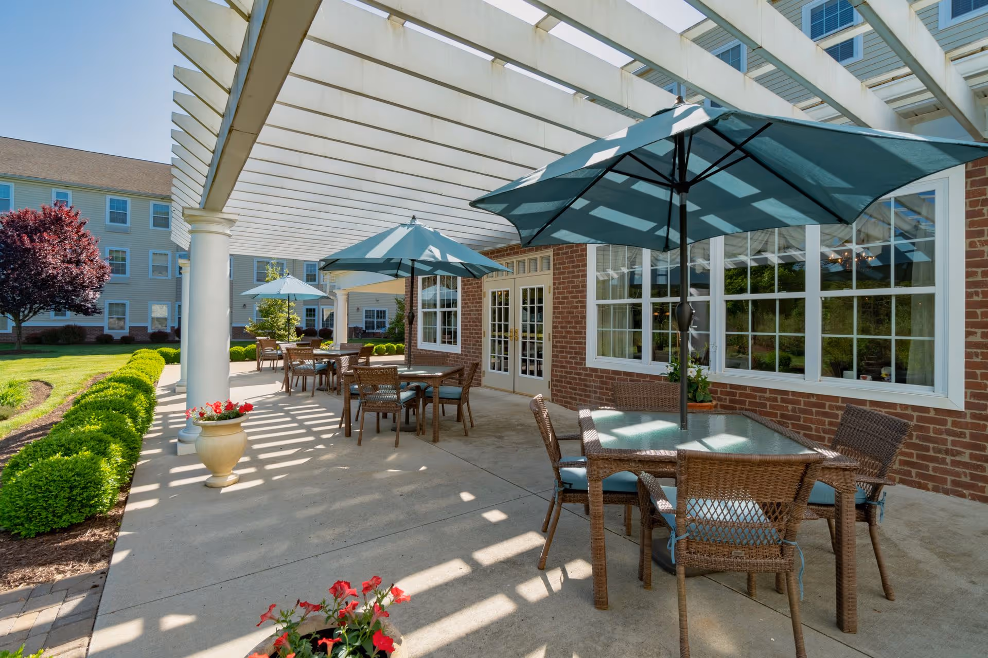 Outdoor patio area at Bentley Commons at Lynchburg with several tables and chairs under large blue umbrellas. The patio is covered by a white pergola casting shadows on the concrete floor. There are manicured bushes and flowers along the edge of the patio, and a brick building with large windows and double doors in the background.