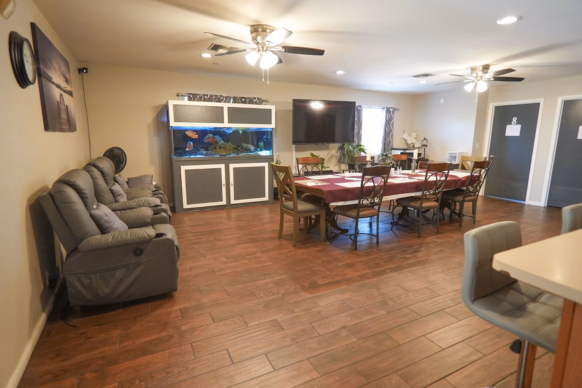 A spacious room with a long dining table covered with a maroon tablecloth and surrounded by chairs. To the left, there are several gray recliner chairs lined up against the wall. A large fish tank is positioned against the far wall beneath a mounted flat-screen TV. The room has wooden flooring, two ceiling fans with lights, and two doors labeled with numbers in the background.