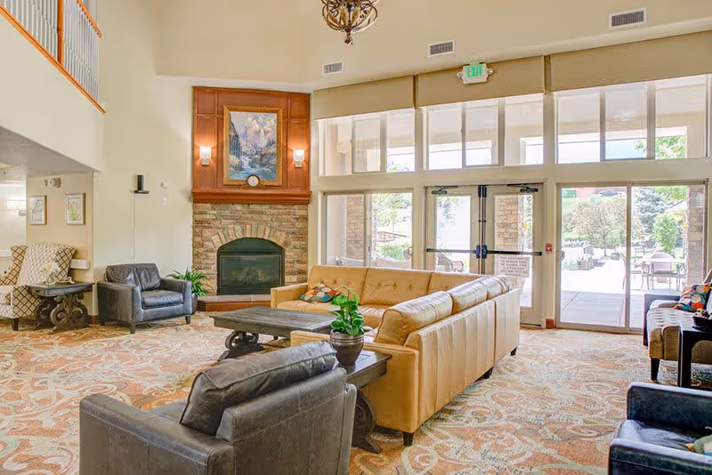 A spacious senior living facility common area with a high ceiling, large windows, and glass doors leading outside. The room features a stone fireplace with a wooden mantle, a large beige sectional sofa, several armchairs, a coffee table with a potted plant, and patterned carpet flooring. The space is well-lit with natural light and has a warm, inviting atmosphere.