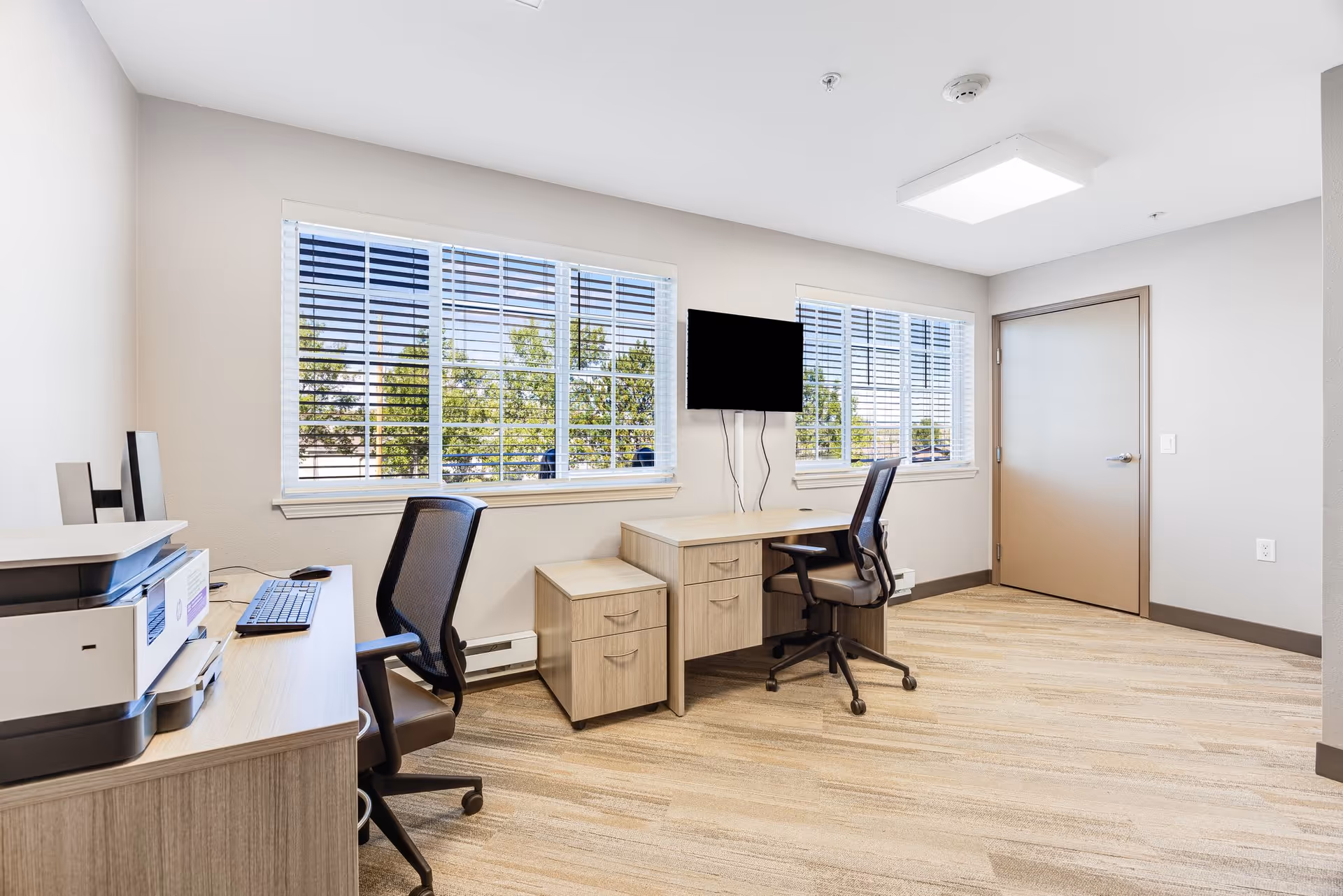 A bright office room with two desks and two office chairs. One desk has a printer and keyboard, while the other has a mounted flat-screen TV above it. Large windows with blinds let in natural light, and a closed door is visible on the right side. The room has light-colored walls and carpeted flooring.