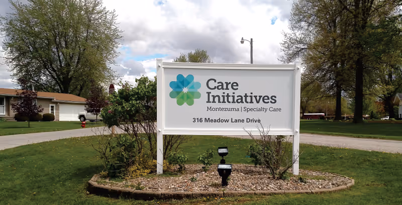 White freestanding sign for Care Initiatives Montezuma Specialty Care on a grassy lawn with trees and houses in the background.