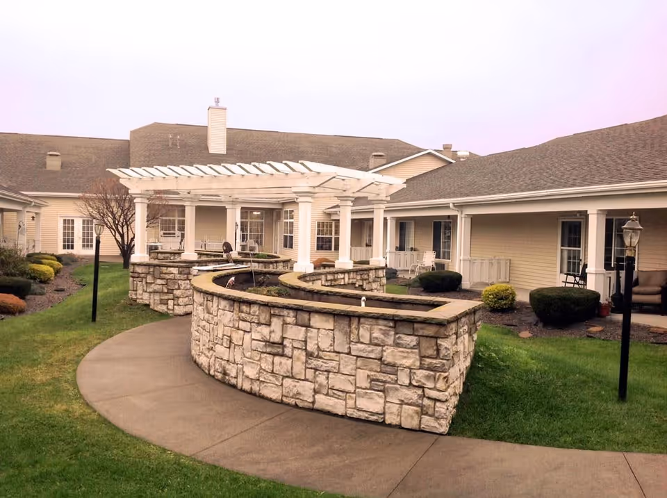 Outdoor courtyard area of a senior living community featuring a curved stone planter with a white pergola above it, surrounded by a paved walkway, green grass, shrubs, and beige buildings with multiple windows and porches.