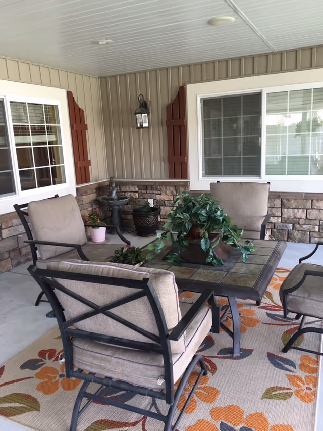 Covered outdoor patio area with four cushioned chairs around a square table with a potted plant centerpiece. The patio has a floral patterned rug, stone wall accents, two windows with white frames, and a wall-mounted lantern light fixture.