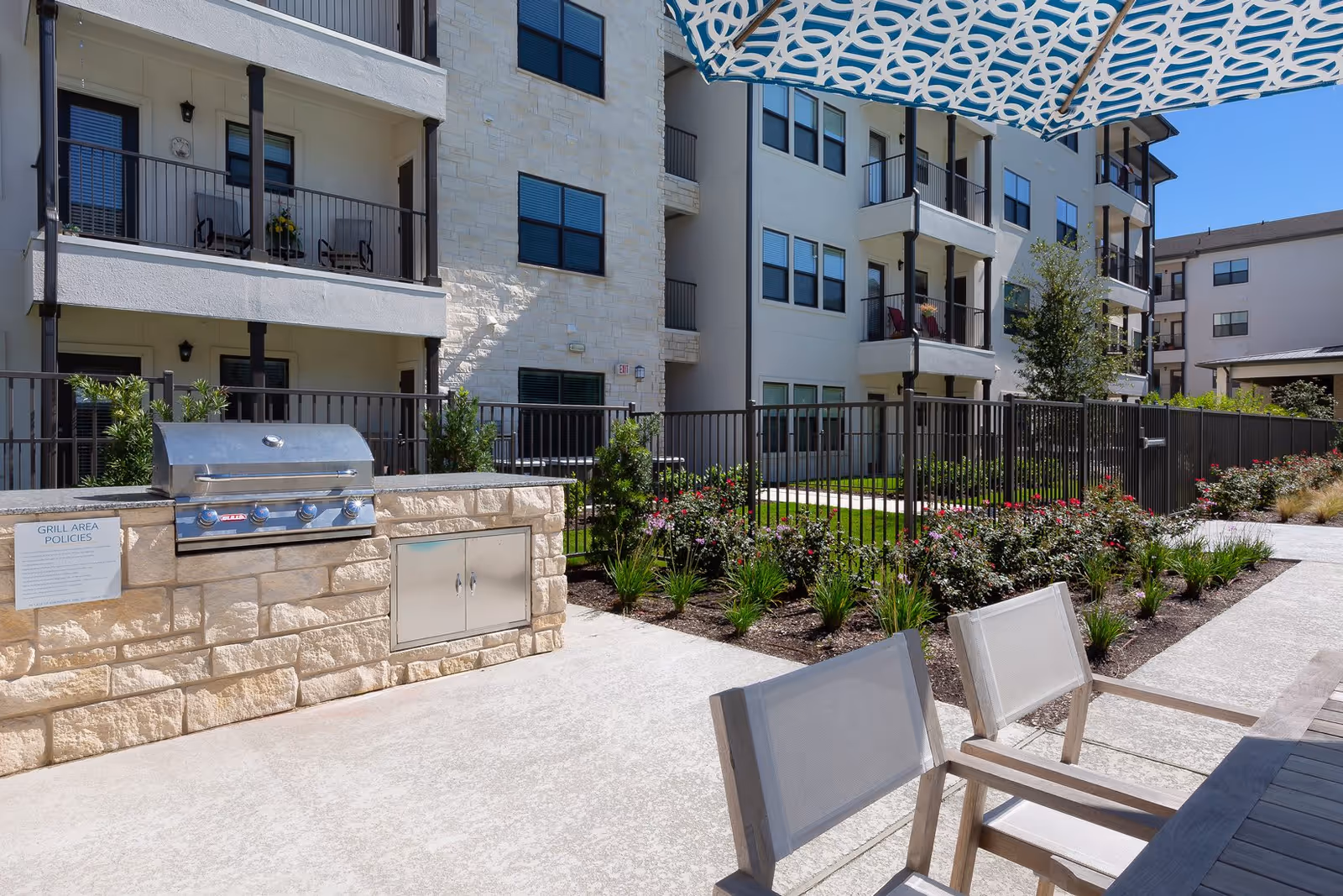 Outdoor patio area at Affinity at Cedar Park featuring a built-in stainless steel grill set in a stone counter, a table with chairs under a patterned umbrella, and a fenced garden with flowering plants. The background shows a multi-story residential building with balconies.
