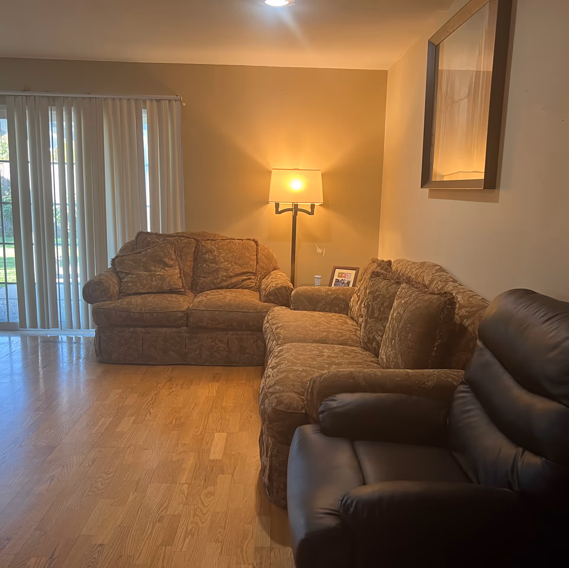 A cozy living room with two patterned brown sofas and a black leather recliner chair. A floor lamp with a warm light is placed between the sofas, and a large mirror hangs on the wall above one sofa. Vertical blinds cover a sliding glass door that leads to an outdoor area with green grass.