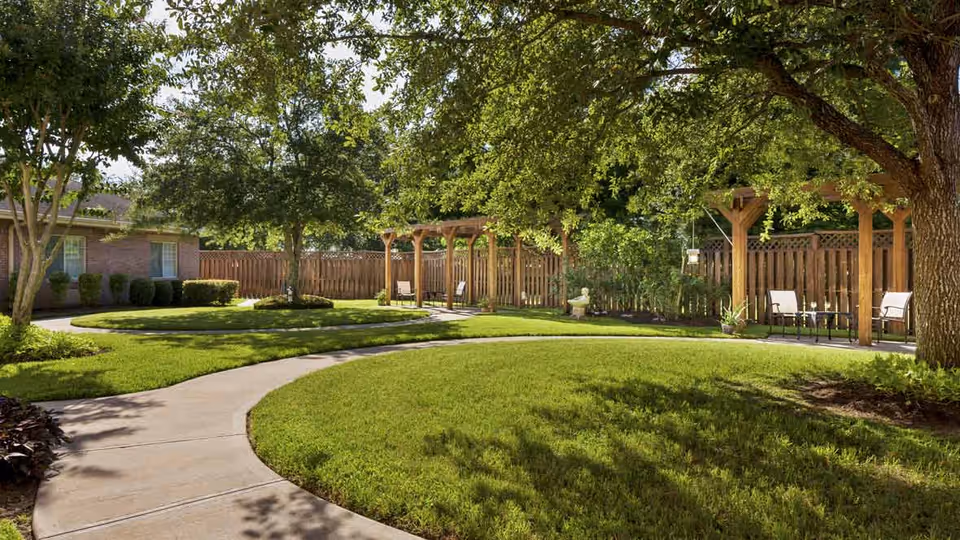 A well-maintained outdoor garden area with a curved concrete walkway, green grass, large trees providing shade, wooden pergolas with seating, and a wooden privacy fence surrounding the space.