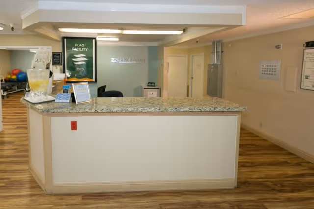 Reception desk area with a granite countertop in Shoreline Healthcare Center. Behind the desk is a glass wall with the Shoreline logo, a green banner on the left side, and some office chairs. The floor has wood-patterned flooring and the walls are light-colored.
