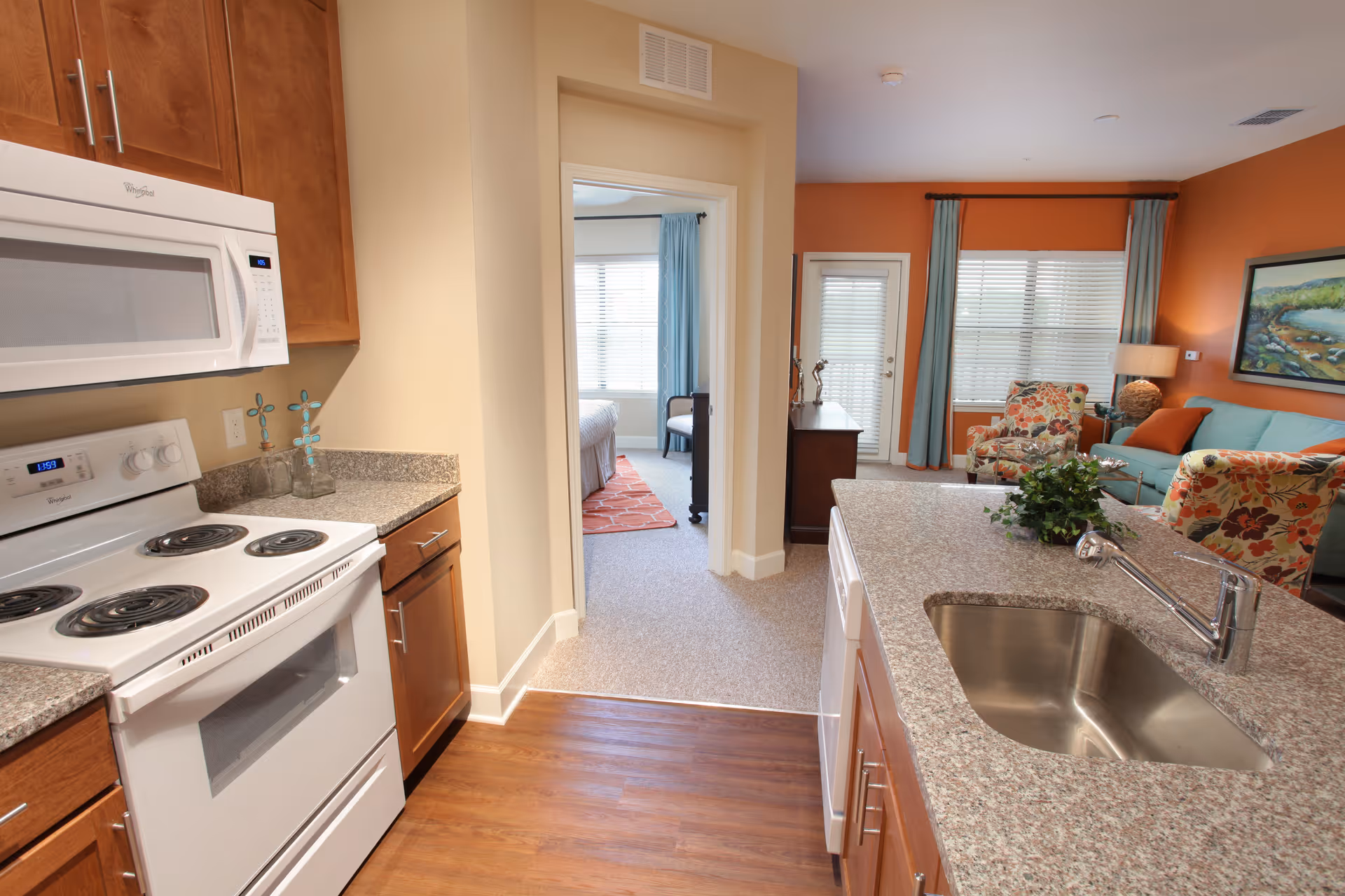 Open-concept apartment interior with a kitchen island and sink in the foreground leading into a living room and bedroom.