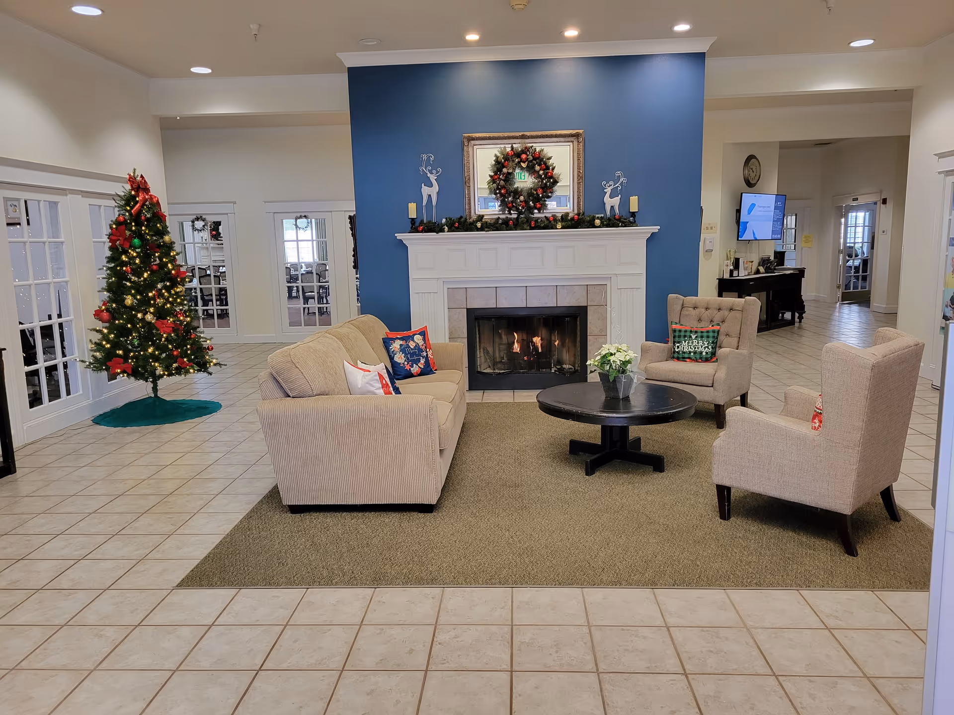 A cozy living room area decorated for Christmas with a lit fireplace, a Christmas wreath and garland on the mantel, and a decorated Christmas tree with lights and red bows. The seating area includes a beige sofa and two beige armchairs with festive pillows around a round black coffee table with a small plant. The room has tiled floors and white walls with a blue accent wall behind the fireplace.