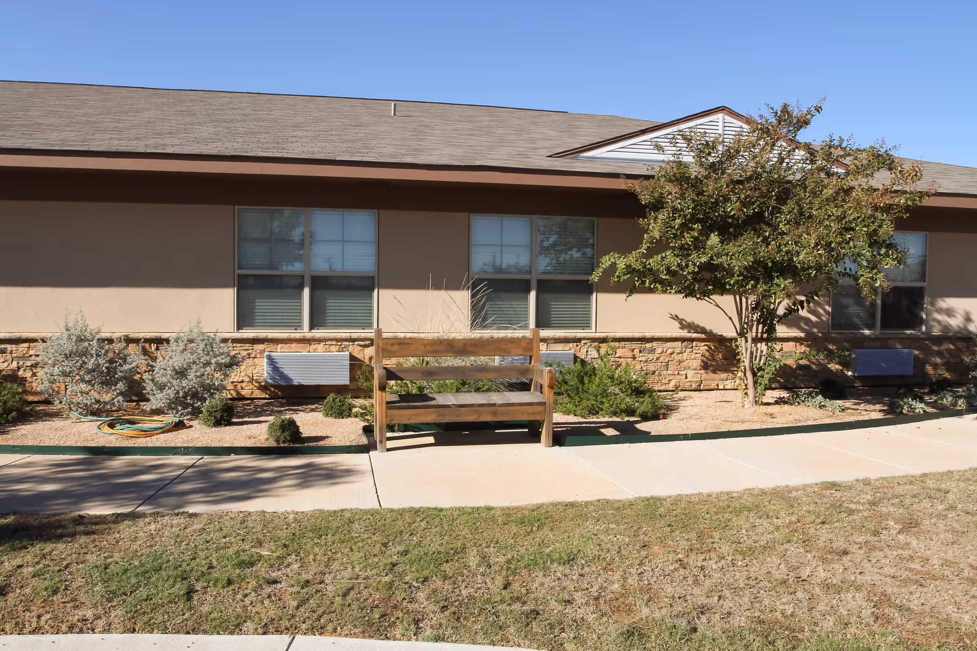 Exterior view of a single-story building with beige walls and stone accents. There are three windows with blinds, a wooden bench on a concrete walkway, some shrubs, a tree, and a garden hose coiled on the ground. The sky is clear and blue.