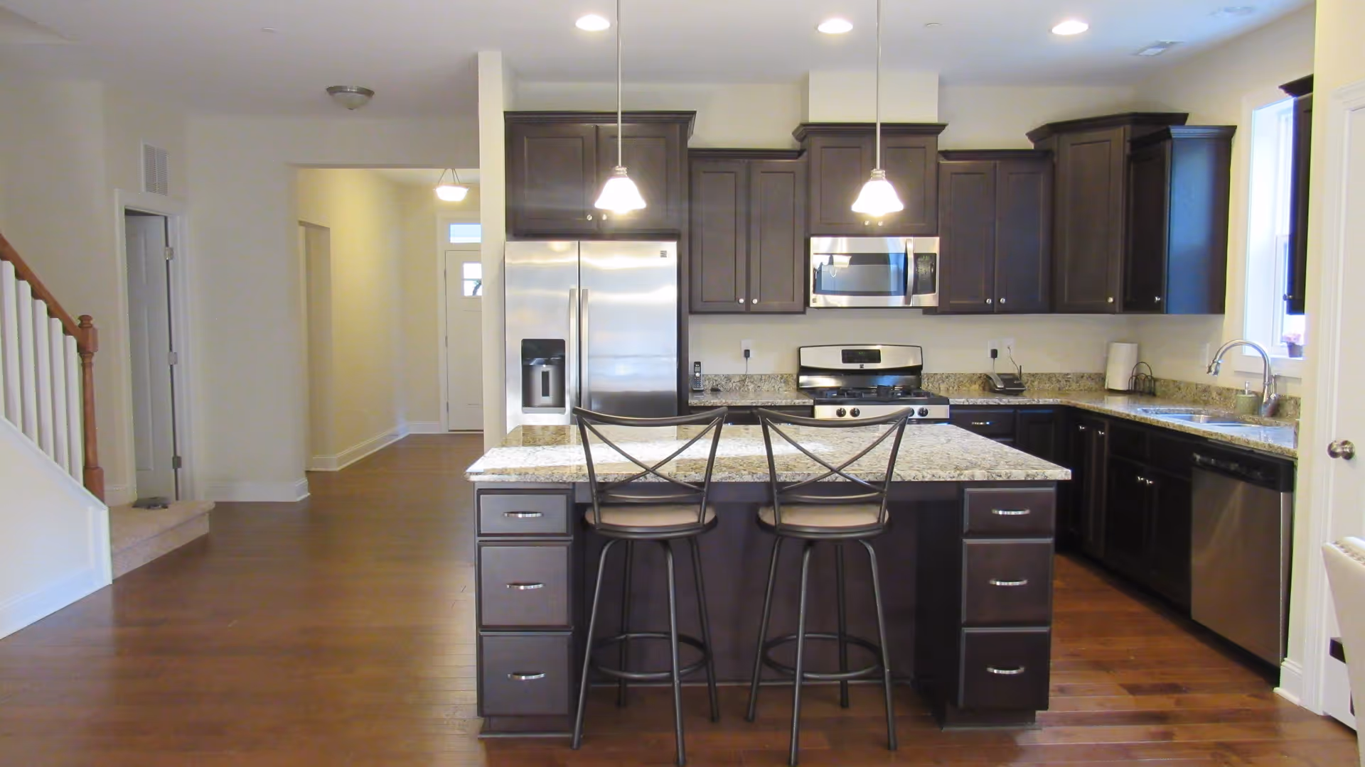 Modern open kitchen with a central island, two bar stools, dark cabinets, stainless steel appliances, and hardwood floors.
