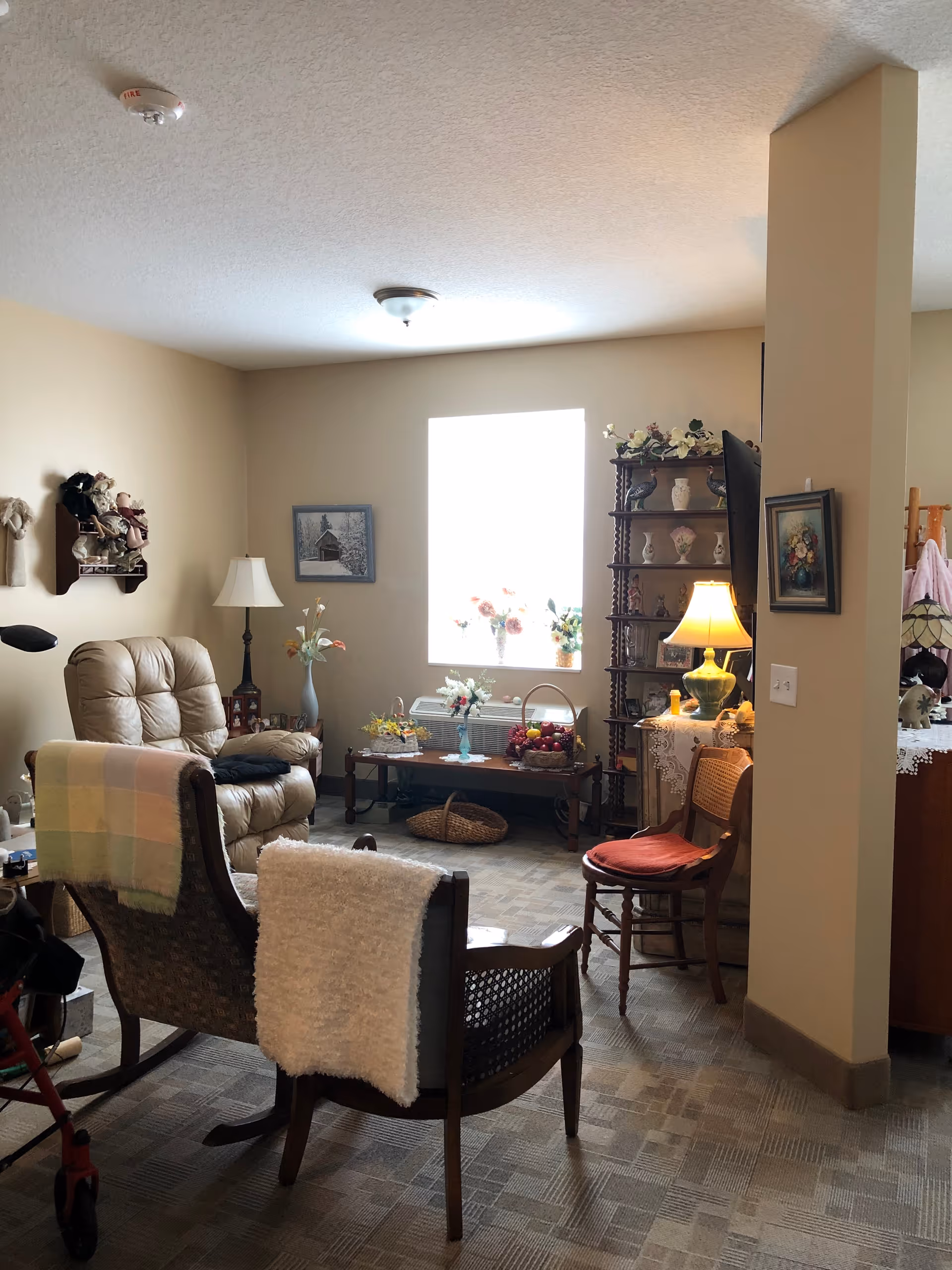 A cozy living room area with beige walls and carpeted floor. The room features a beige recliner chair, a wooden rocking chair with a white blanket draped over it, and a wooden chair with a red cushion. There is a wooden shelf filled with decorative items and a table with a lamp and more decorations. A window lets in natural light, illuminating flower arrangements on a table beneath it. The room has a warm and inviting atmosphere with various homey decorations.