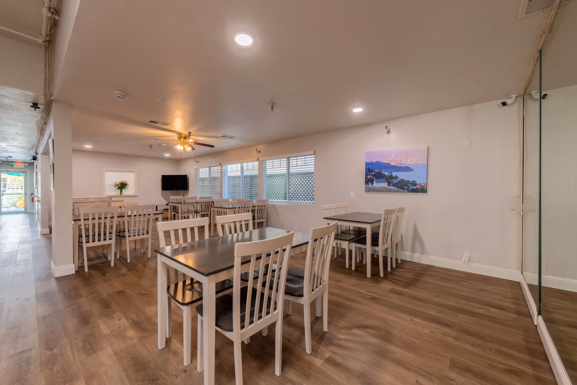 A dining room with multiple tables and chairs arranged neatly on a wooden floor. The room has white walls, a ceiling fan with lights, a wall-mounted TV, and a large window with blinds. There is a framed picture on the wall and a glass partition on the right side.