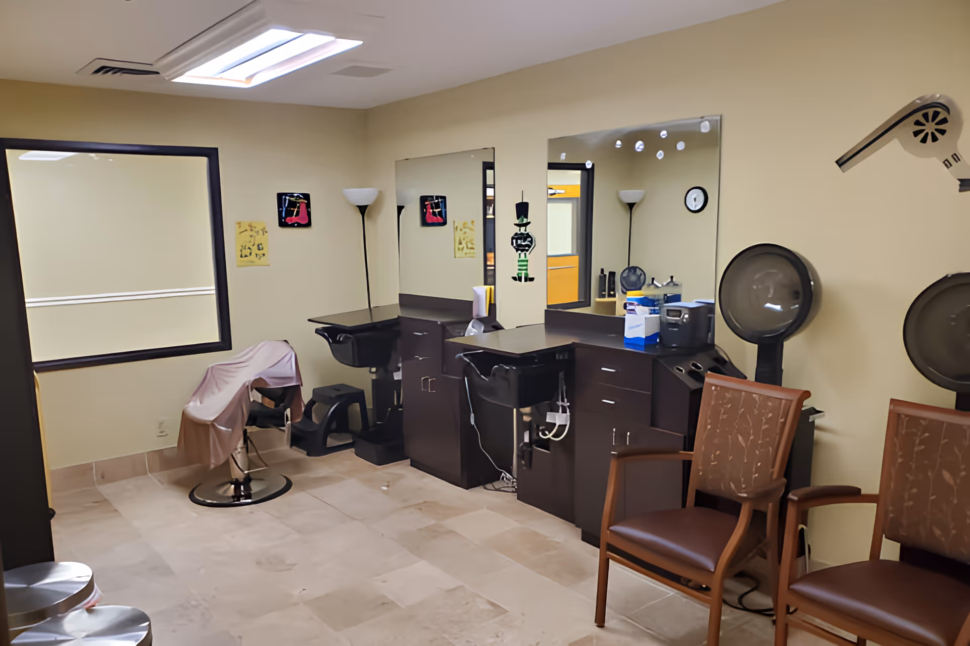 Interior view of a hair salon area in a senior living facility with salon chairs, mirrors, hair dryers, and cabinets. The room has beige walls and tiled flooring, with two brown chairs on the right side and a large window on the left.