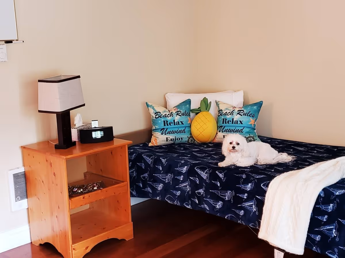 A cozy bedroom corner with a wooden nightstand holding a lamp, tissue box, and clock radio. Next to it is a bed with a navy blue sailboat-patterned bedspread, two decorative pillows with the text 'Beach Rules Relax Unwind Enjoy,' a pineapple-shaped pillow, and a small white dog lying on the bed. A white knitted blanket is draped over the edge of the bed.