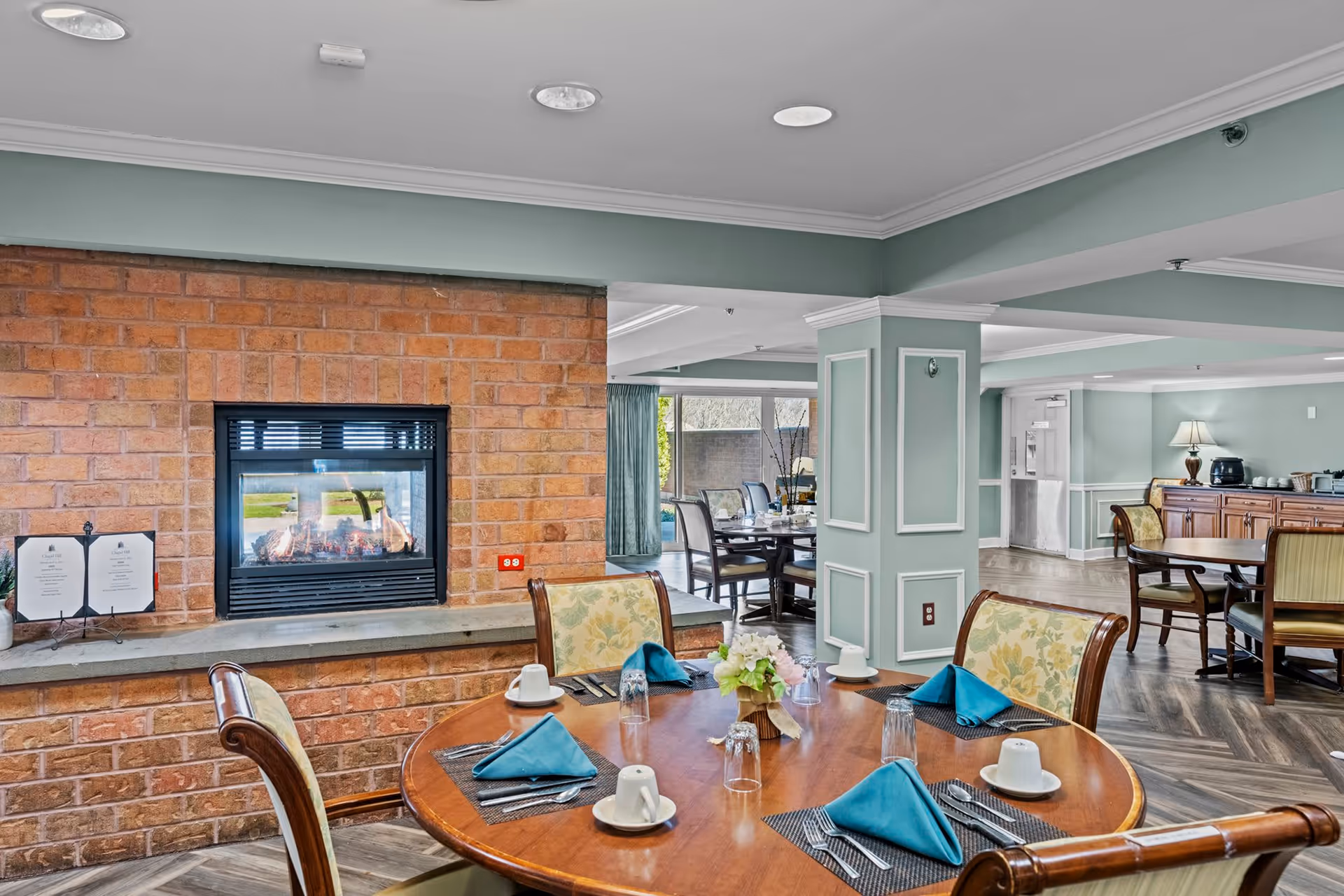 Dining area in a senior living facility with a round wooden table set with blue napkins, cups, and utensils. The room features a brick fireplace, light green walls with white trim, wooden chairs with floral upholstery, and large windows letting in natural light.