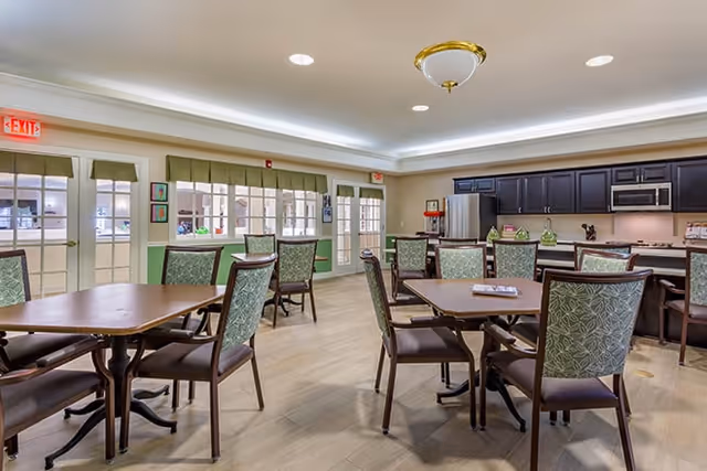 A bright and spacious dining area with several wooden tables and chairs featuring green patterned upholstery. The room has large windows with green valances, light-colored walls, and a kitchen area with dark cabinets, a stainless steel refrigerator, and a microwave. The floor is light wood, and there are exit signs above the doors.