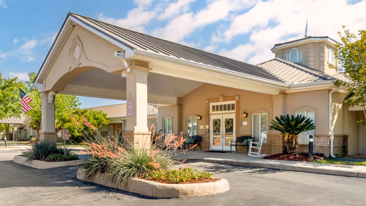 Entrance of Brookdale Oakwell senior living facility with a covered drop-off area, benches, rocking chairs, landscaped plants, and an American flag under a partly cloudy sky.