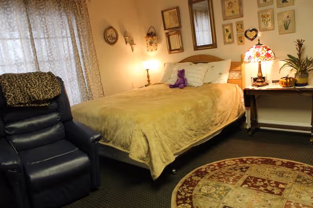 Cozy bedroom with a made bed topped by a purple stuffed bear, a side table with a lamp, a recliner chair, and a round decorative rug.