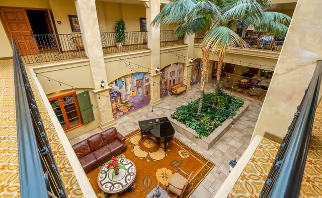 Upper-level view of a multi-story indoor atrium with a grand piano, seating area, large potted palms, and decorative murals.