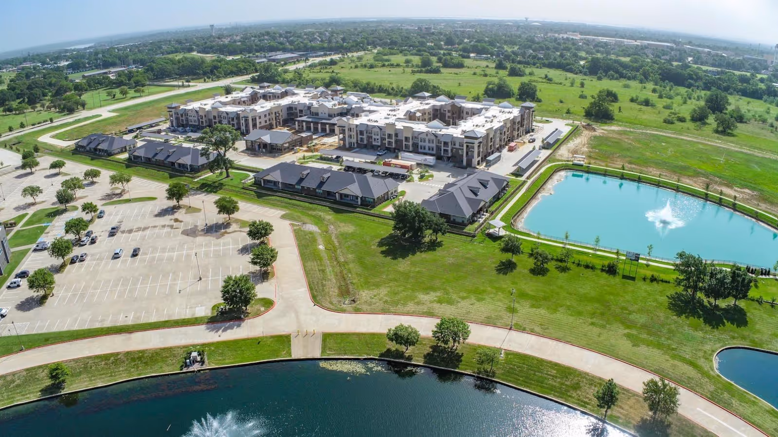 Aerial view of Watermere at Flower Mound senior living facility showing multiple buildings, parking lots, green lawns, walking paths, and two ponds with fountains, surrounded by open fields and trees.